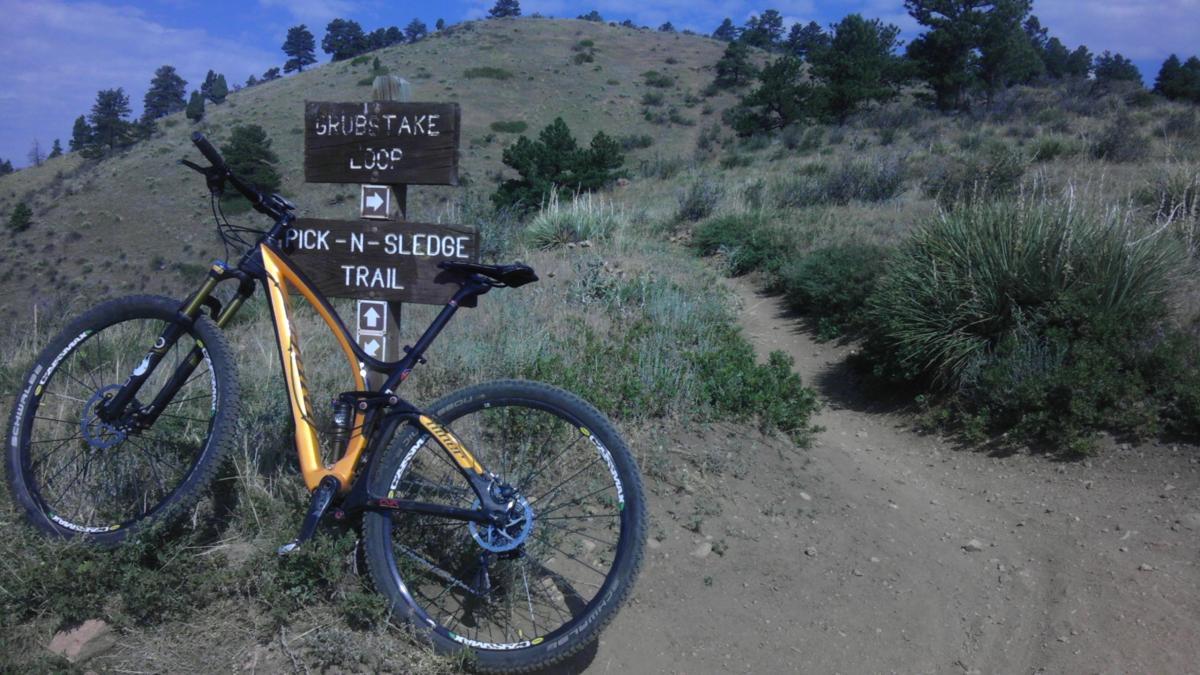 A mountain bike leaning against a wooden trail sign with directional arrows, indicating the paths for "Grubstake Loop" and "Pick-N-Sledge Trail," set against a backdrop of rolling hills and sparse vegetation under a blue sky. Apex Park mountain bike trail.