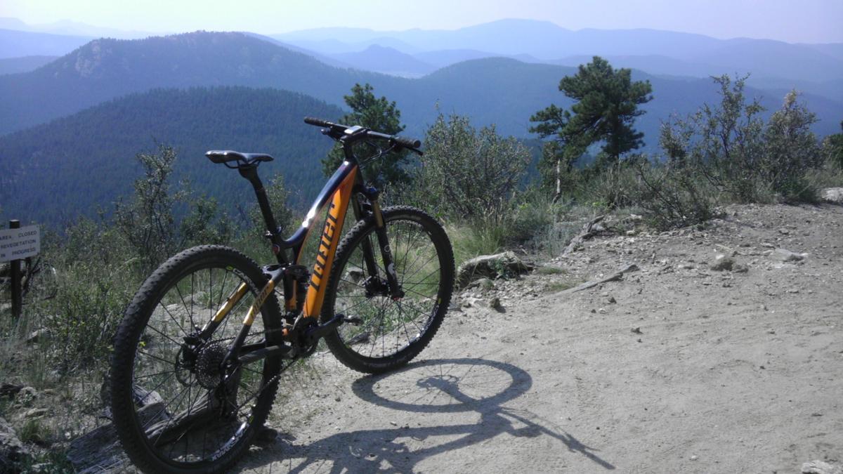 A mountain bike resting on a dirt path, overlooking a scenic view of rolling hills and forests in the background. The landscape features various shades of green with a hazy blue sky and distant mountains. Bergen Peak mountain bike trail.