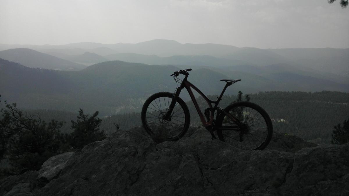 A mountain bike perched on a rocky outcrop, overlooking a misty landscape of rolling hills and mountains in the distance. The scene captures a sense of adventure and the beauty of nature. Bergen Peak mountain bike trail.