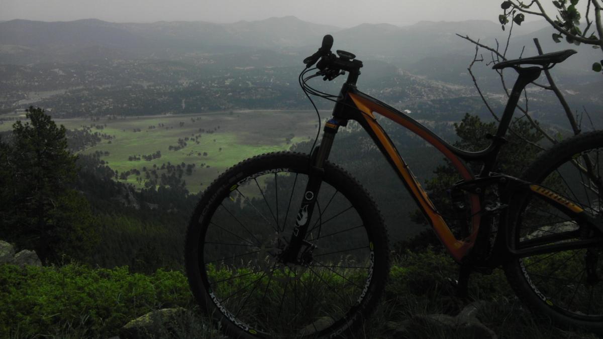A mountain bike positioned on a rocky outcrop overlooking a valley with green fields and distant mountains under a cloudy sky. Bergen Peak mountain bike trail.