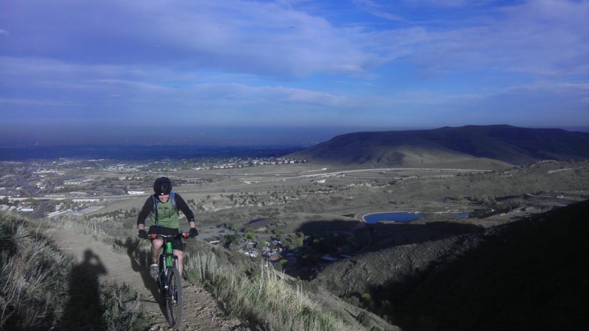 A mountain biker rides along a narrow trail with a scenic view of a valley below, surrounded by rolling hills and a blue sky with scattered clouds. The landscape features green shrubs, residential areas, and a small pond in the distance. Apex Park mountain bike trail.
