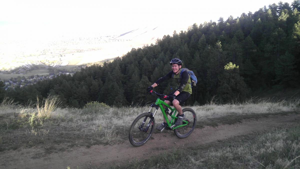 A person riding a green mountain bike on a dirt trail, surrounded by trees and hills, with a scenic view of a valley in the background. The rider is wearing a helmet and a backpack, smiling as they navigate the trail. Apex Park mountain bike trail.