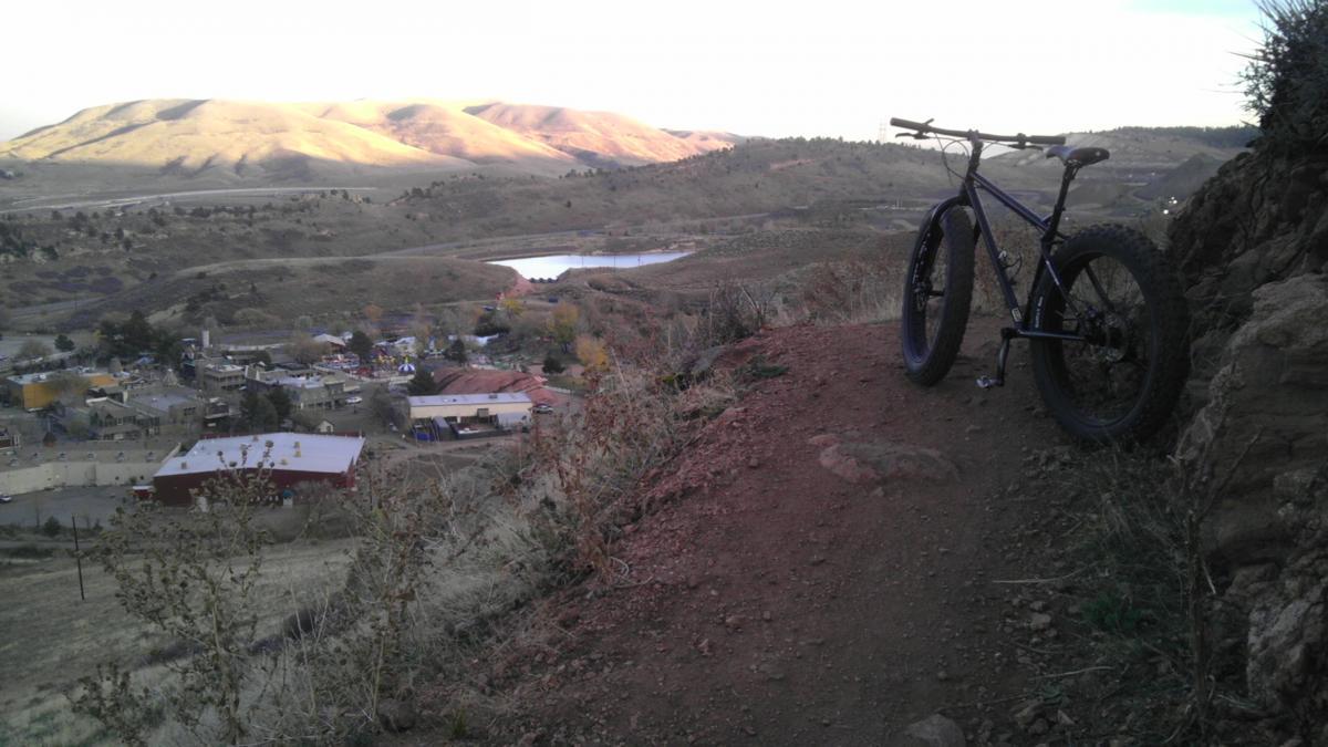 A fat-tire mountain bike is parked on a rocky ledge overlooking a picturesque valley, with hills in the background and a small town nestled below. A winding river can be seen reflecting the light, surrounded by dry vegetation and autumn-colored trees. The scene captures the beauty of outdoor biking in a serene landscape. Apex Park mountain bike trail.
