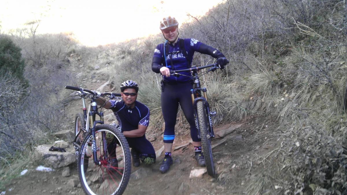 Two mountain bikers pose on a narrow dirt trail surrounded by dry grass and rocky terrain. One biker is squatting next to his bike, while the other stands beside his bike, both wearing helmets and athletic clothing appropriate for biking. The background shows a hilly landscape under bright sunlight. Apex Park mountain bike trail.