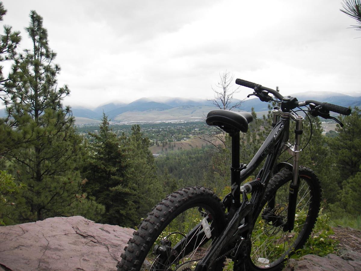 A mountain bike leaning against a rock, overlooking a scenic view of a valley surrounded by trees and mountains under a cloudy sky. Blue Mountain mountain bike trail.