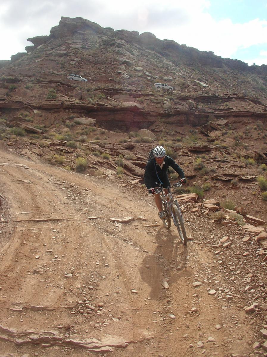 A cyclist wearing a helmet rides down a rocky dirt trail with a steep hill in the background. Sparse vegetation is visible among the rocky terrain, and a couple of vehicles are parked on a higher section of the trail. The sky is partly cloudy, suggesting an outdoor adventure in a natural setting. White Rim Trail mountain bike trail.