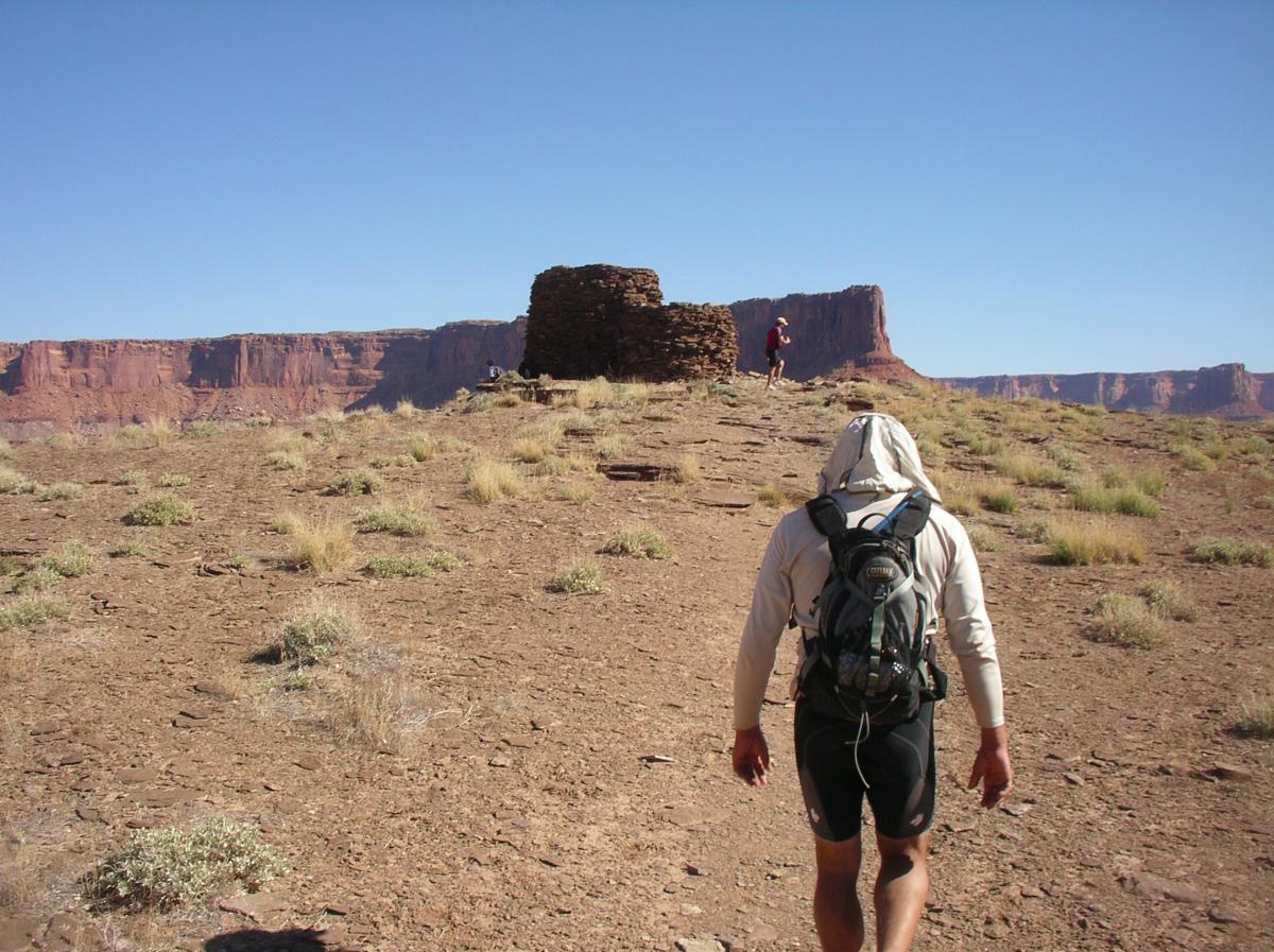 A hiker wearing a light-colored hoodie and a backpack walks toward a stone structure on a barren, grassy landscape, with red rock cliffs in the background under a clear blue sky. Another individual is seen in the distance near the structure. White Rim Trail mountain bike trail.