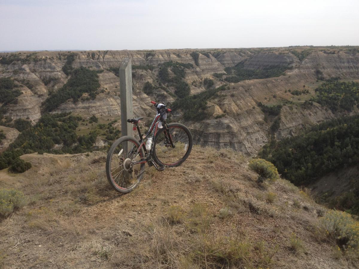 A mountain bike leaning against a wooden post overlooking a rugged canyon landscape. The scene features steep, layered rock formations and patches of greenery in the canyon below, with a cloudy sky overhead. Maah Daah Hey mountain bike trail.