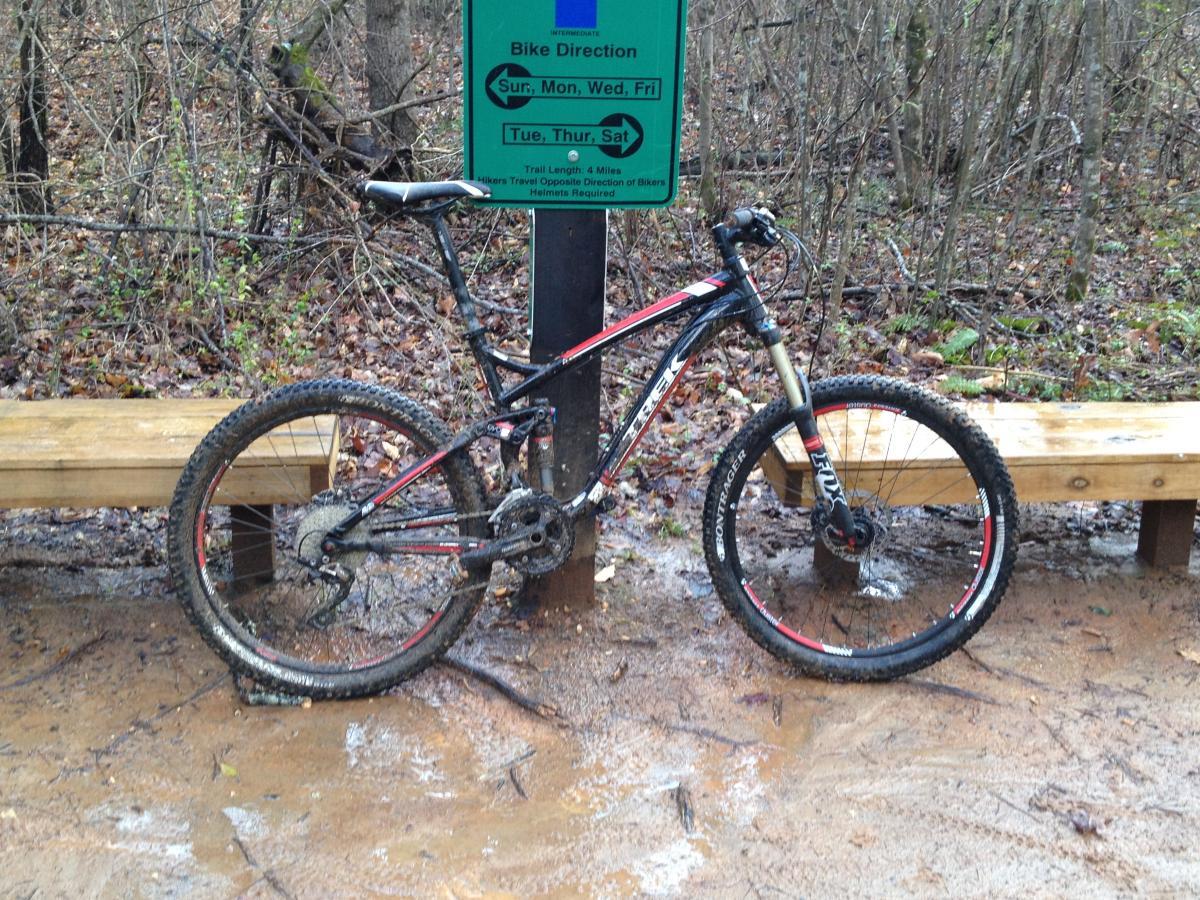 Trek Fuel EX 7: A mountain bike covered in mud is leaning against a trail sign. The sign indicates bike direction and includes information about trail lengths and days for biking. A wooden bench is visible in the foreground, and the surrounding area is forested with wet, muddy ground and fallen leaves.