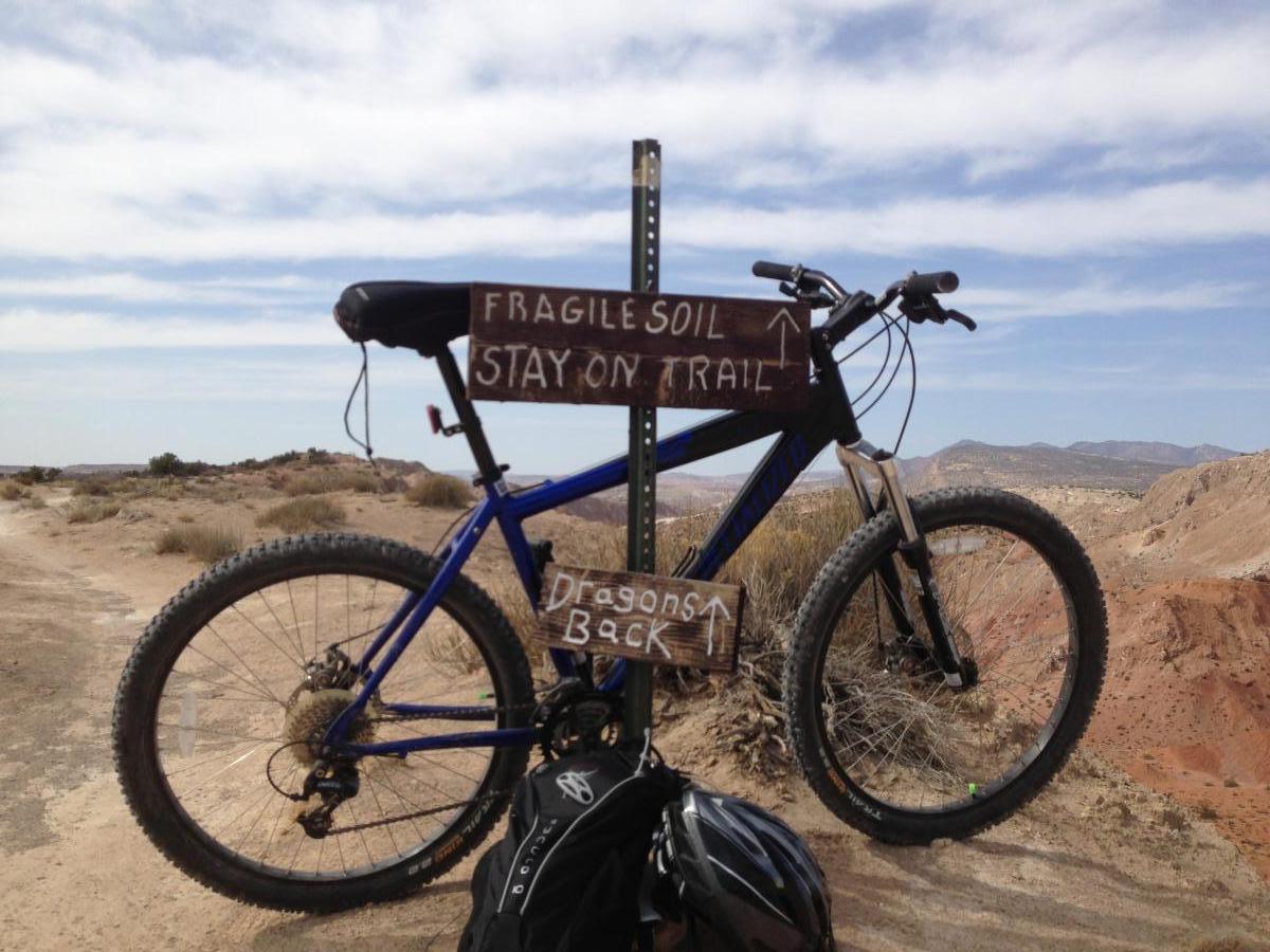 A blue mountain bike resting against a signpost in a desert landscape. The sign has wooden plaques indicating "FRAGILE SOIL" and "STAY ON TRAIL," with an arrow pointing upward. A backpack and helmet are leaning against the bike. The background features arid terrain and distant mountains under a blue sky with white clouds. White Ridge Bike Trails mountain bike trail.