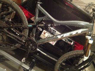 Yeti ASR 5 Alloy: A muddy mountain bike mounted on a bike rack, positioned in a garage with a black vehicle in the background. The bike is angled to show its frame and wheels, with visible dirt on the tires and components.