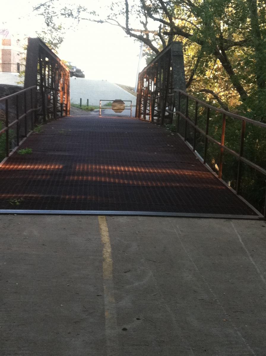 A rusted metal bridge leads toward a closed barrier, with a tree-lined path beside it. The sunlight casts shadows on the bridge's surface, and a faint outline of a flat area is visible in the background, indicating a road or pathway ahead. Spadra Creek Nature Trail mountain bike trail.