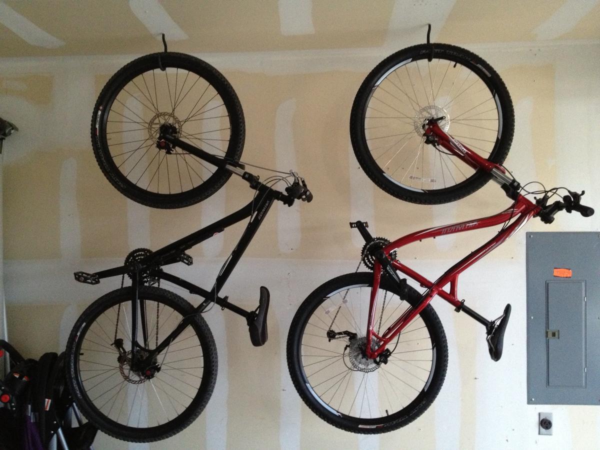 Specialized Myka Disc: Two mountain bikes hanging from the wall in a garage. The bike on the left is black, and the bike on the right is red. The garage walls are unfinished, with visible drywall patterns. A power panel is also visible in the background.