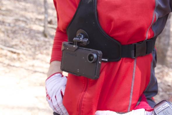 A person wearing a red long-sleeve shirt with a chest harness mounted with an action camera, standing outdoors in a forested area. The action camera is attached to the harness, positioned at the person's chest.