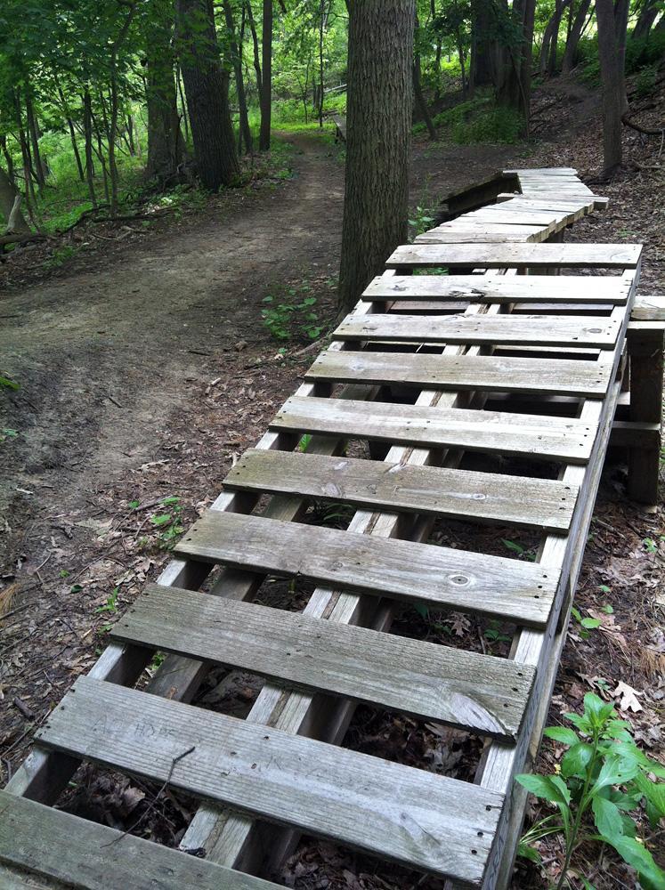 A wooden plank bridge winding through a forested area, with a dirt path visible to the side. The bridge has slatted wooden boards and is surrounded by green foliage and trees. Jermain Park mountain bike trail.