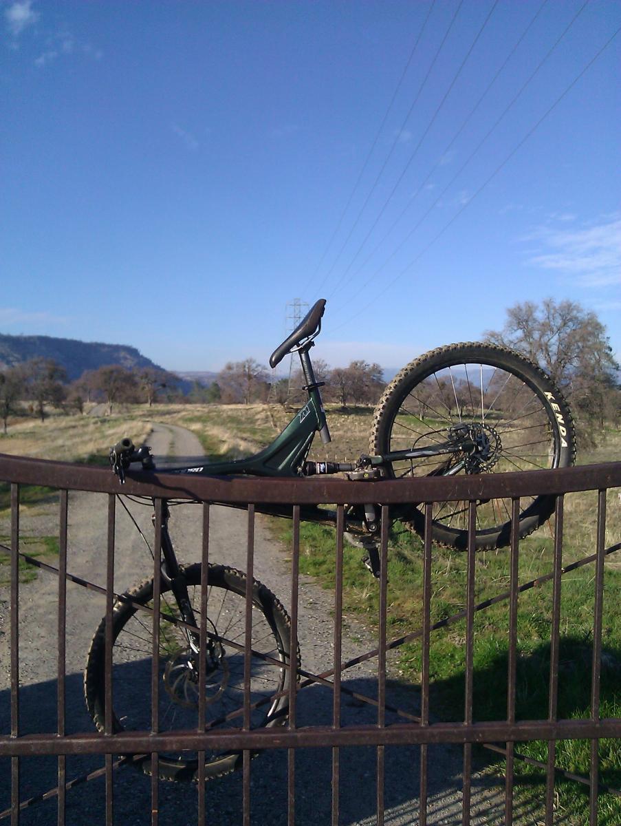 Specialized Enduro Expert: A mountain bike resting against a metal gate, with a dirt path and grassy fields in the background under a clear blue sky. Power lines run across the scene, and hills are visible in the distance.