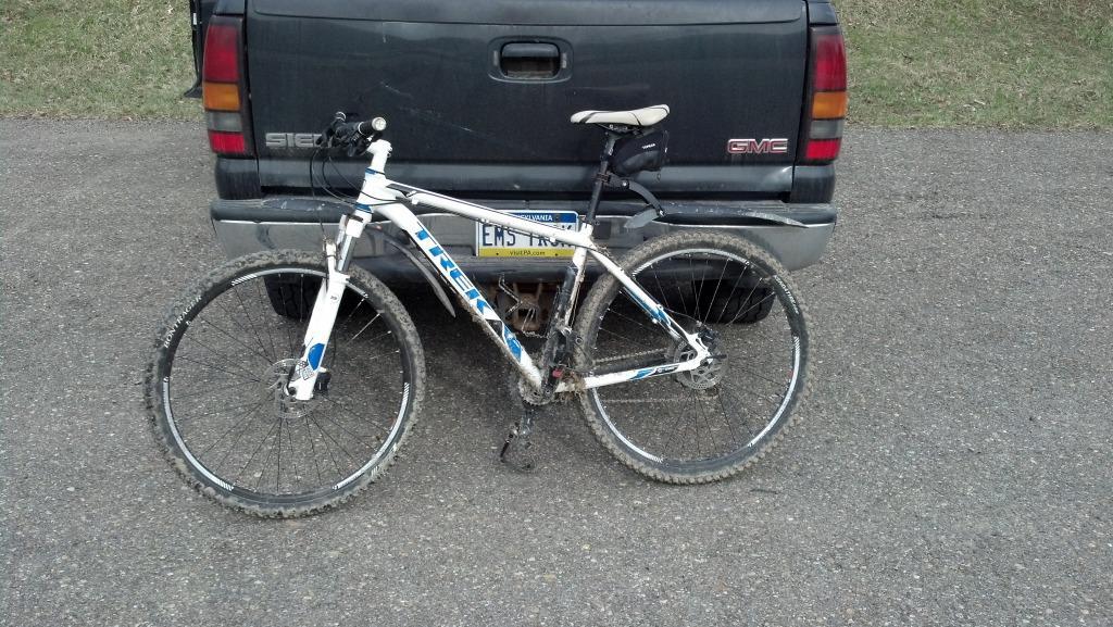 Trek Mamba: A mountain bike with a white and blue frame, leaning against the back of a black GMC pickup truck on a gravel surface. The bike appears to have some dirt on it, indicating recent use. The truck's rear features a license plate, partially visible, and the area is surrounded by grass.