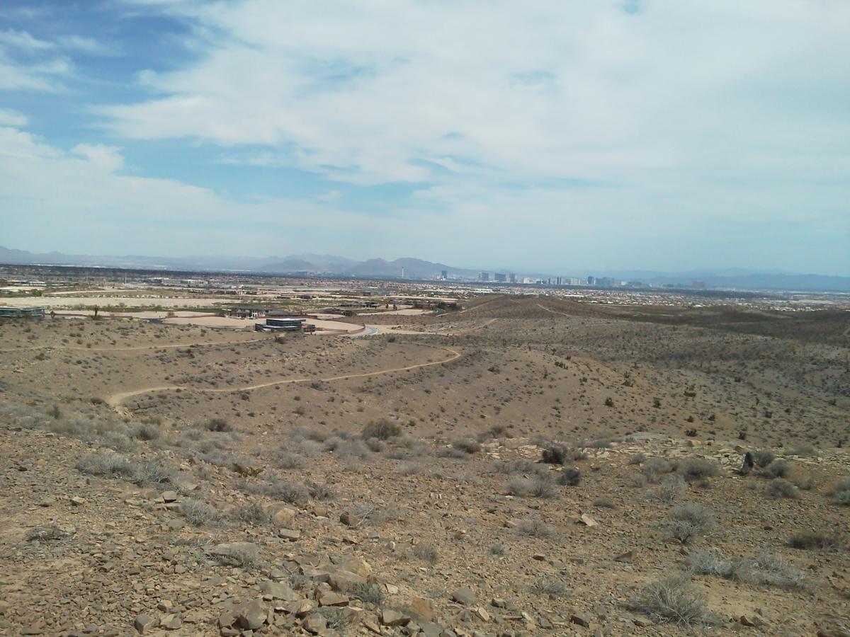 A panoramic view of a desert landscape with rocky terrain, sparse vegetation, and a winding dirt path. In the distance, the skyline of a city is visible beneath a partly cloudy sky, with mountains framing the horizon. Bears Best mountain bike trail.