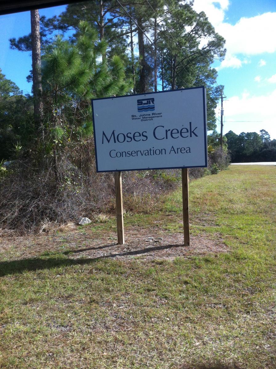 Sign for Moses Creek Conservation Area, featuring the logo of the St. Johns River Water Management District. Surrounded by greenery and trees, with a grassy area in the foreground and a clear sky above. Moses Creek mountain bike trail.