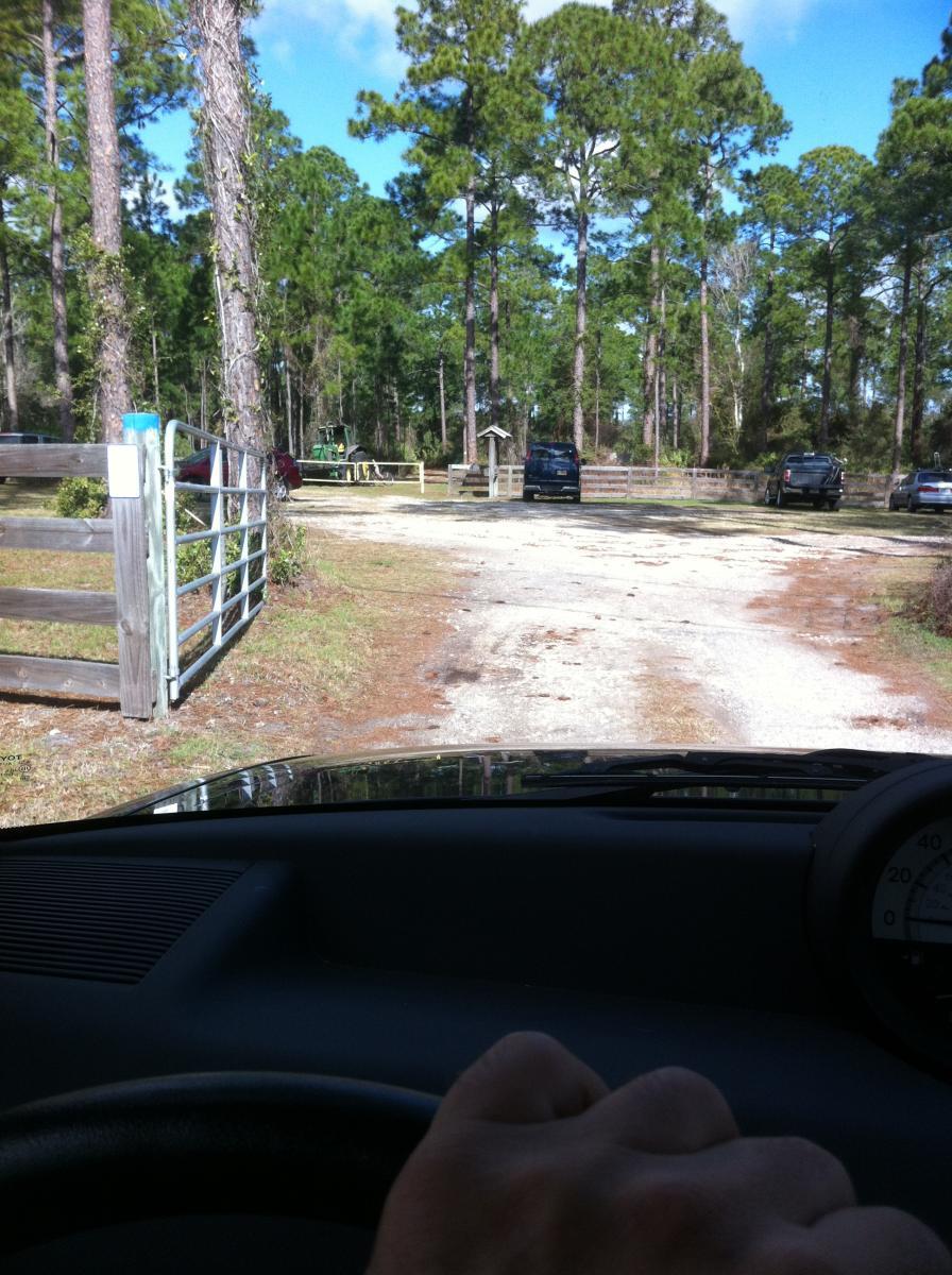 A view from inside a vehicle showing a gravel road leading through a wooded area with tall pine trees. A metal gate is visible on the left, and several parked vehicles can be seen in the background. The sky is partly cloudy, indicating a sunny day. Moses Creek mountain bike trail.