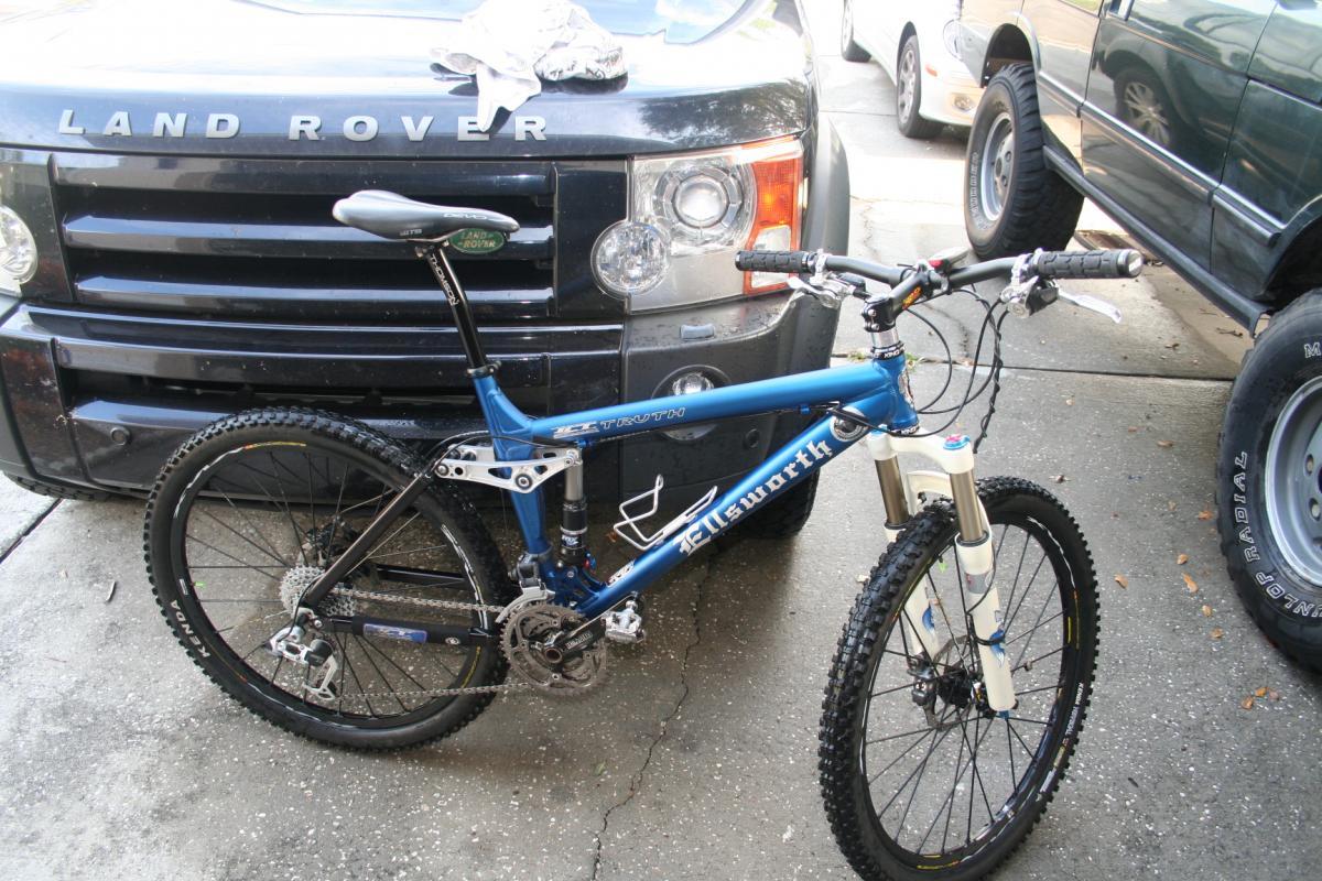 Ellsworth Truth: A blue mountain bike is parked next to a black Land Rover, showcasing its suspension and rugged tires. The foreground features the bike’s sleek frame and components, while the vehicle's front grille is prominently visible in the background. The setting appears to be a driveway, with light reflections on the car and a clear surface underneath.