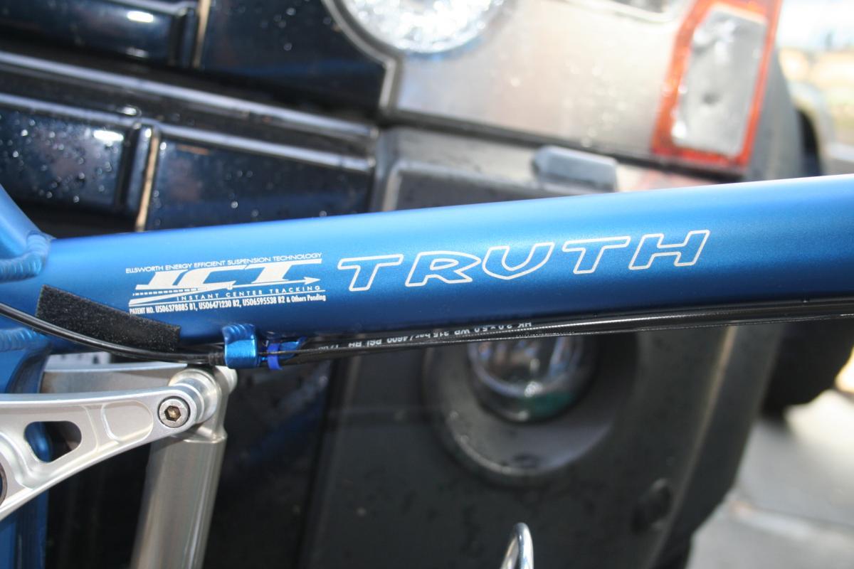 Close-up view of a blue bicycle frame displaying the word "TRUTH" in bold letters, with details of the frame's suspension technology and components in the background. A blurred vehicle is partially visible, suggesting the bicycle is parked near it.