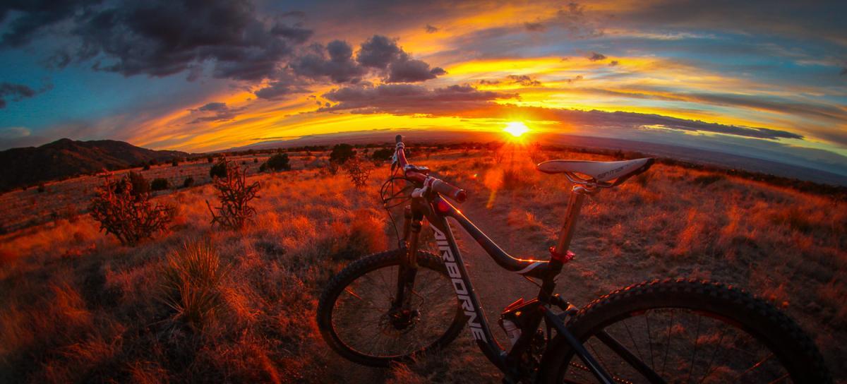 A mountain bike parked on a trail with a vibrant sunset in the background, featuring orange, pink, and purple hues in the sky, along with scattered clouds and distant mountains. The foreground is filled with golden grass and shrubs, creating a serene outdoor scene. South Foothills mountain bike trail.