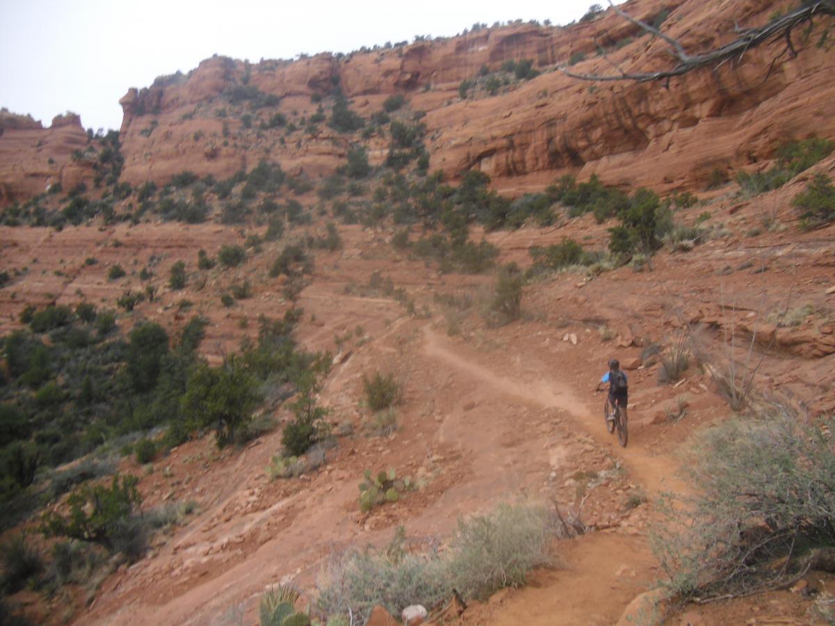 A person riding a mountain bike on a dirt trail through a rugged, rocky landscape with reddish-orange cliffs and sparse vegetation in the background. Mescal Trail mountain bike trail.