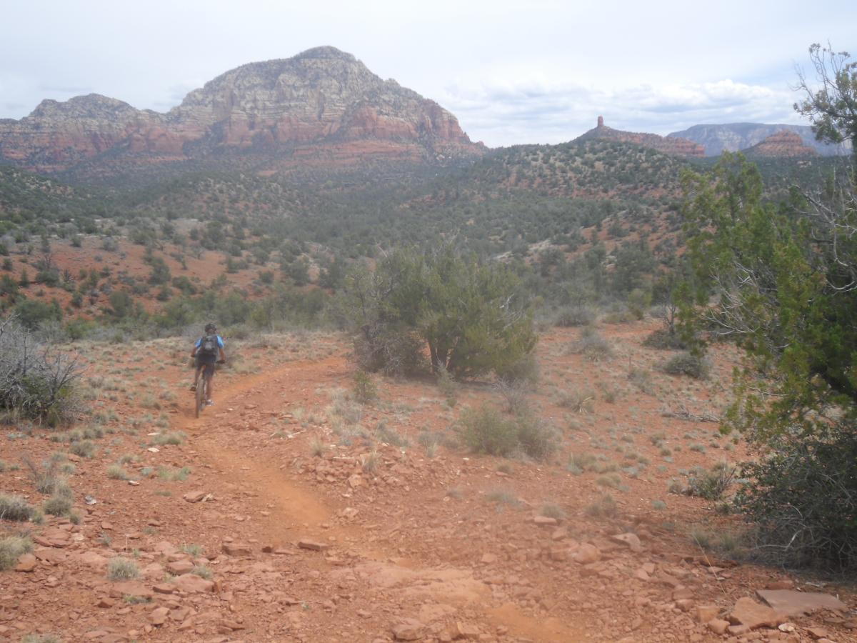 A person riding a mountain bike on a dirt trail surrounded by red rock formations and vegetation in a rugged landscape. The scene features distant mountains under a cloudy sky, showcasing natural beauty and outdoor adventure. Mescal Trail mountain bike trail.