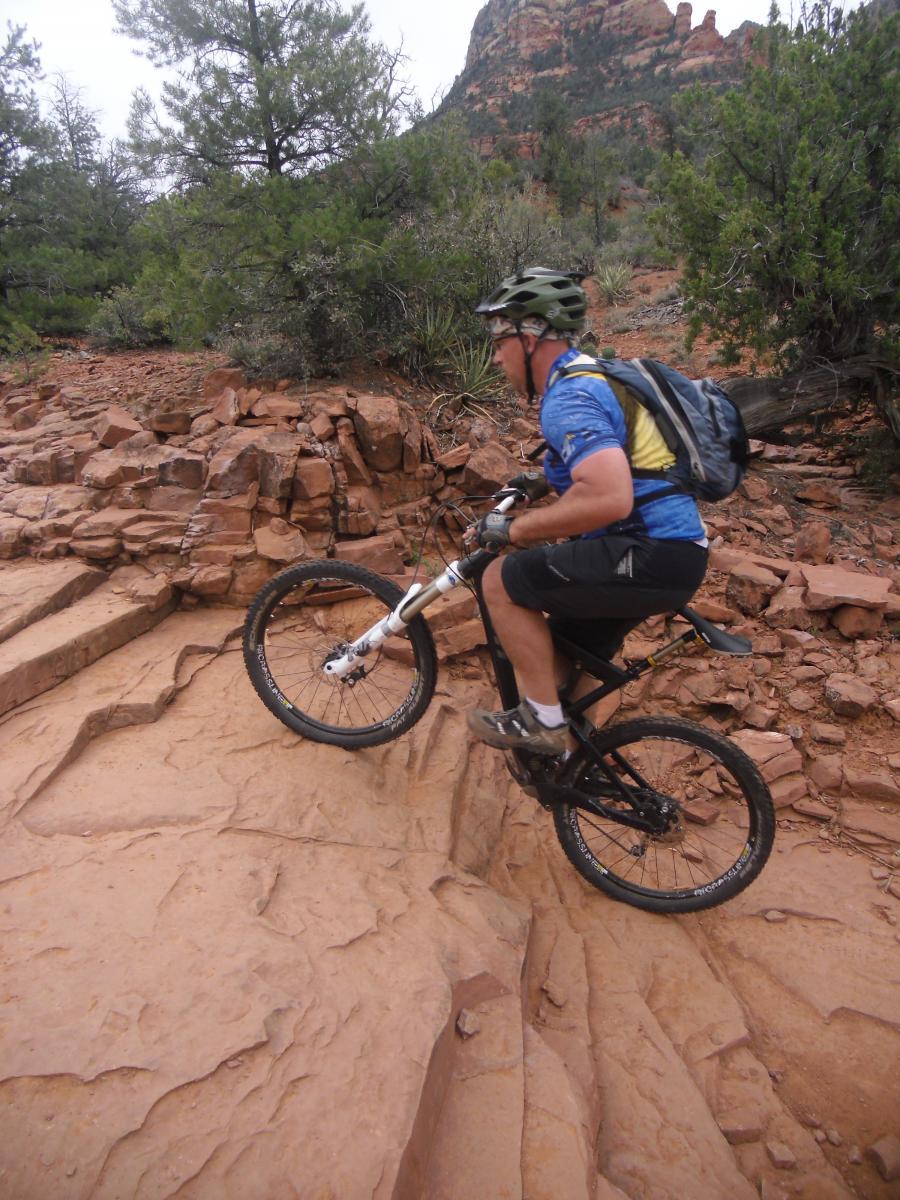 A mountain biker navigating a rocky trail in a desert landscape, with red rock formations and greenery in the background. The cyclist is wearing a helmet and a blue and yellow jersey, demonstrating skill as he maneuvers over uneven terrain. Chuck Wagon mountain bike trail.