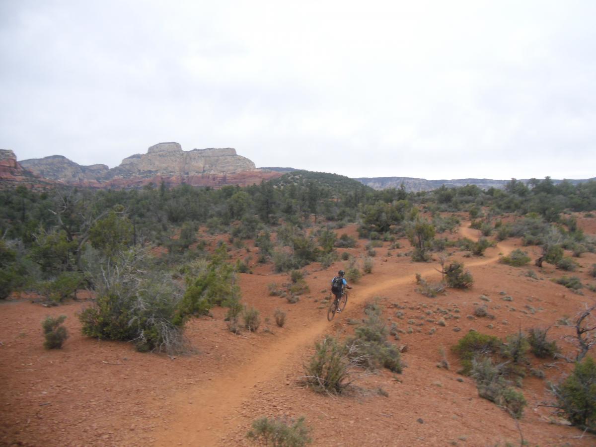 A person riding a mountain bike along a winding dirt trail in a desert landscape, with red soil and scattered shrubs, set against rugged rock formations under a cloudy sky. Chuck Wagon mountain bike trail.
