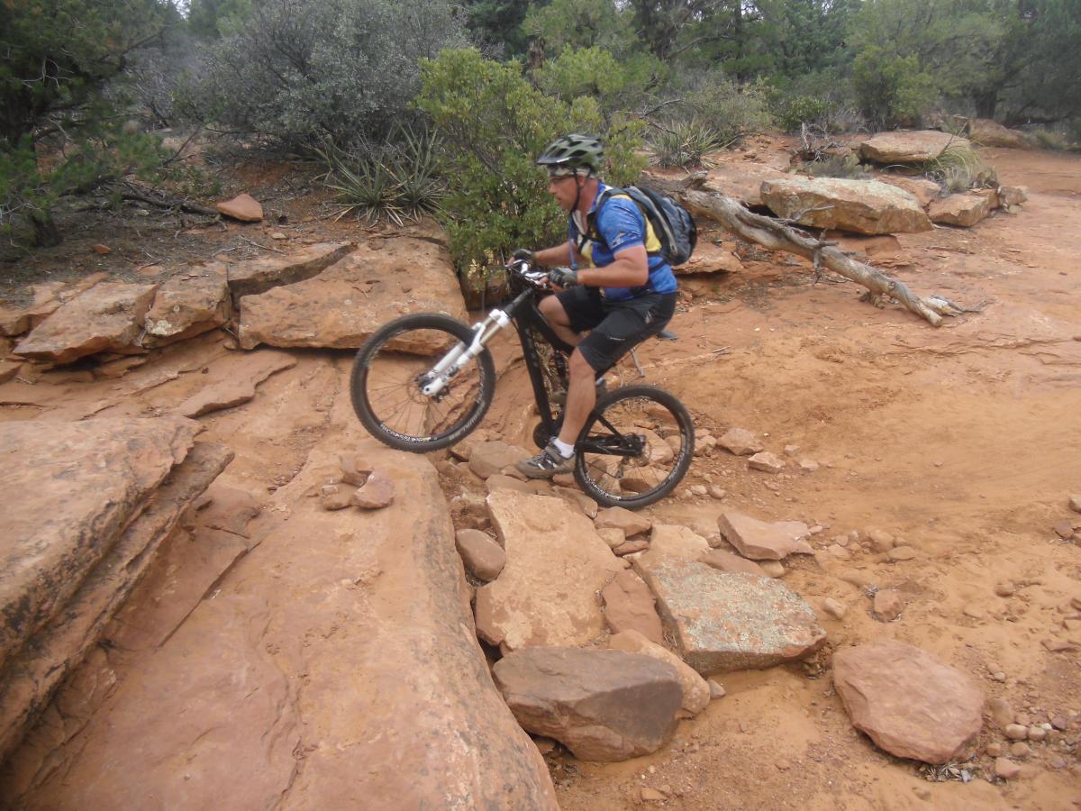 A mountain biker navigating rocky terrain on a dirt trail, surrounded by greenery and natural landscapes. The rider is in a dynamic position, with one wheel lifted over a large rock while wearing a helmet and backpack. Chuck Wagon mountain bike trail.