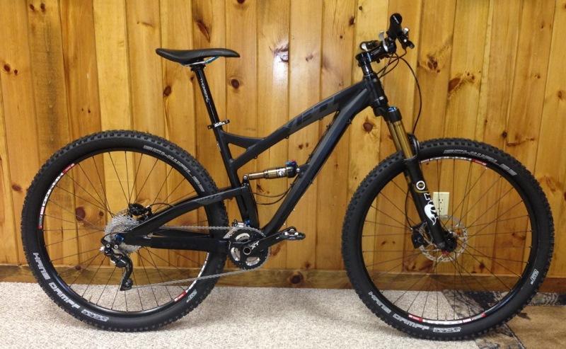 Yeti SB95: A black mountain bike with a sleek design, featuring wider tires, front suspension, and a prominent frame, is positioned against a wooden wall. The bike has disc brakes and is equipped for off-road biking.