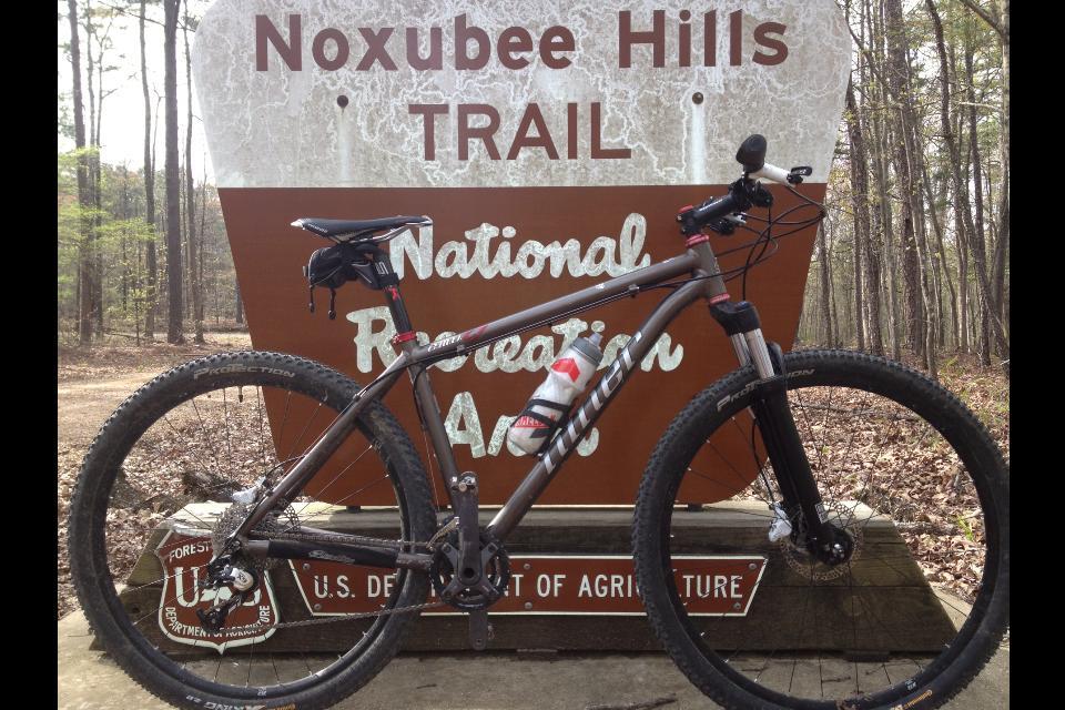 Niner EMD 9: A mountain bike parked next to a "Noxubee Hills Trail" sign in a forested area, with trees in the background and dirt path visible on the ground. The sign displays the words "National Recreation Area" and includes U.S. Department of Agriculture branding.