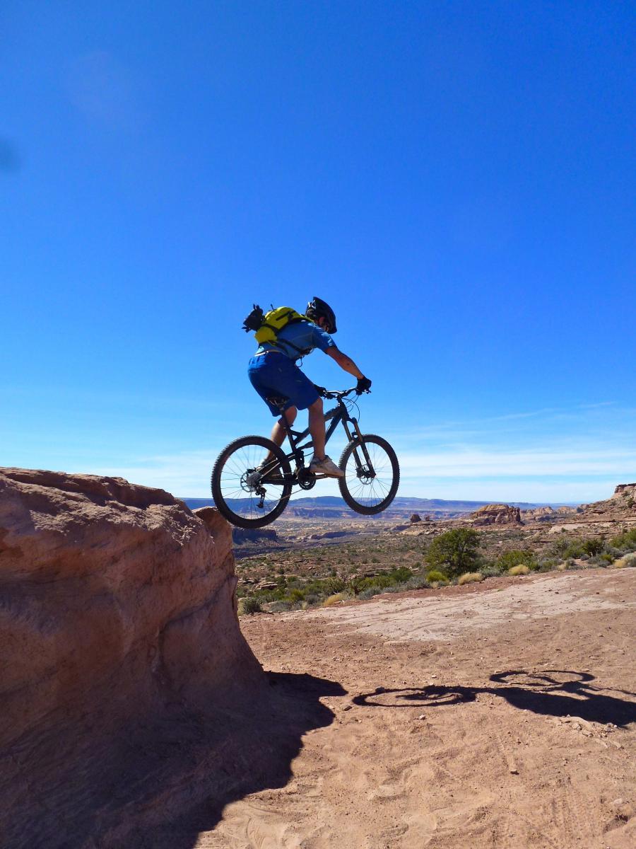 A mountain biker performing a jump over a rocky ledge, with a clear blue sky and a scenic landscape in the background. The rider is wearing a helmet and backpack, showcasing an action-packed moment in outdoor sports. Porcupine Rim mountain bike trail.