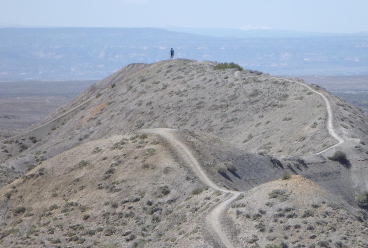 A view of rugged hills with a winding dirt path traversing their slopes. A solitary figure can be seen standing at the top of a hill, surrounded by a vast, open landscape under a clear sky. Zippety Do Dah mountain bike trail.