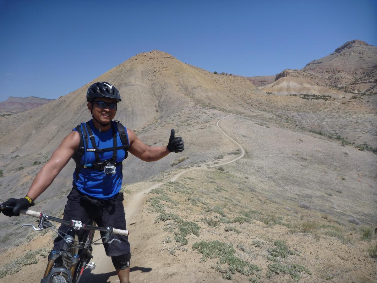 A mountain biker in a blue sleeveless shirt and helmet poses with a thumbs-up while standing next to his bike on a rugged trail. The background features dry, hilly terrain under a clear blue sky. Zippety Do Dah mountain bike trail.