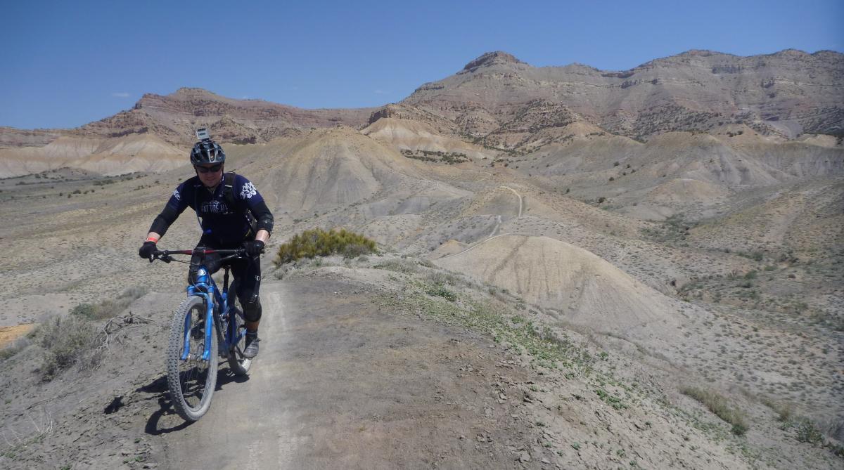 A mountain biker ascends a dirt trail on a rugged terrain with hills in the background. The sun is shining in a clear blue sky, highlighting the varied earthy tones of the landscape, while the biker is equipped with protective gear and a helmet. Zippety Do Dah mountain bike trail.