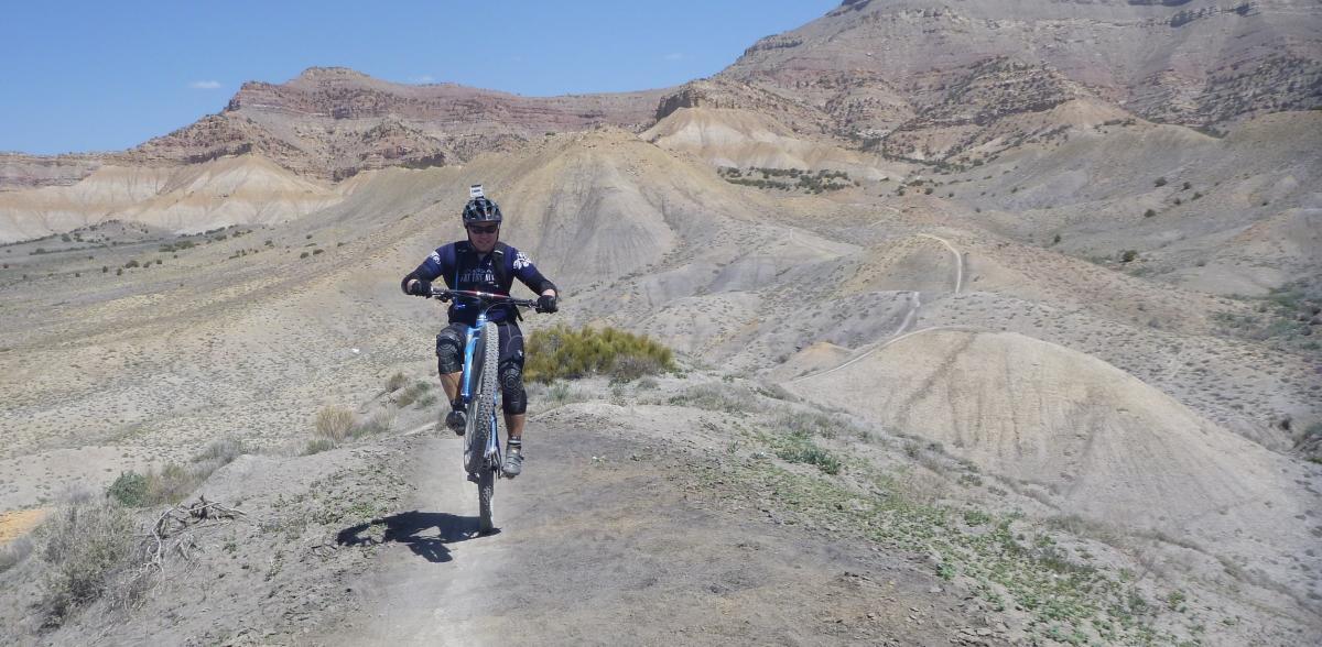 A mountain biker performing a wheelie on a dirt trail with rocky, hilly terrain in the background under a clear blue sky. Zippety Do Dah mountain bike trail.