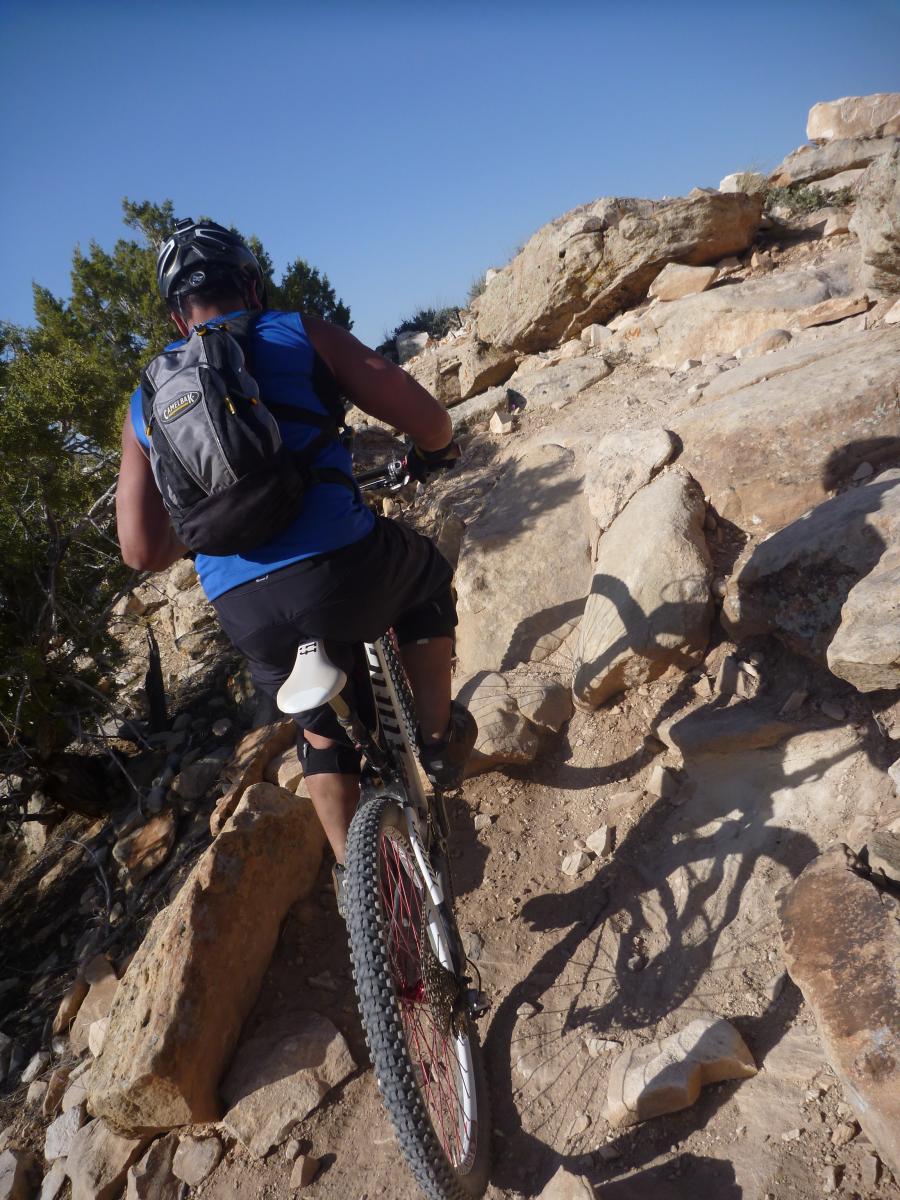 A mountain biker navigates a rocky trail, showcasing a challenging ascent with steep, uneven terrain. The cyclist is wearing a helmet and a blue tank top, with a backpack secured for the ride. Surrounding the path are rugged rocks and sparse vegetation under a clear blue sky. Moore Fun mountain bike trail.
