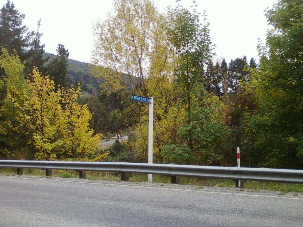 A roadside view featuring a blue street sign that reads "McMillan Rd," surrounded by lush greenery and vibrant autumn foliage. The background shows rolling hills and trees, indicating a natural, rural setting. Moonlight Circuit mountain bike trail.