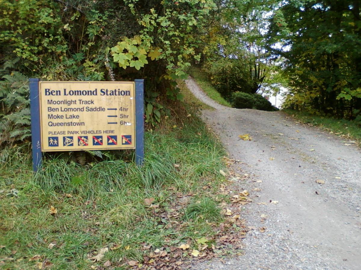 A sign at Ben Lomond Station displaying trail information including the Moonlight Track, Ben Lomond Saddle, Moke Lake, and Queenstown, with estimated hiking durations. The sign is surrounded by lush greenery and indicates parking instructions for vehicles. Moonlight Circuit mountain bike trail.