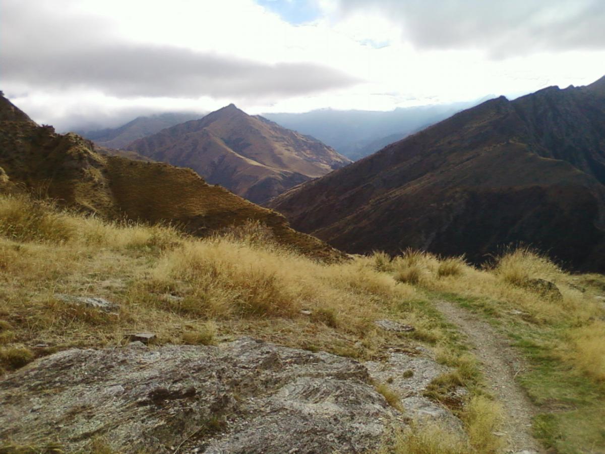 A scenic view of rugged mountains under a cloudy sky, with rolling hills covered in golden grass and rocky terrain leading into the distance. Moonlight Circuit mountain bike trail.