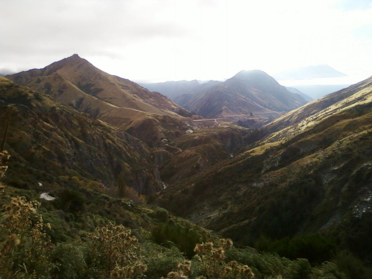 A panoramic view of rugged mountains and valleys, showcasing a mix of green and brown slopes under a cloudy sky. The terrain features steep hills and winding paths, with a small stream visible in the lower foreground. The distant peaks are partially shrouded in mist, adding a mystical quality to the landscape. Moonlight Circuit mountain bike trail.