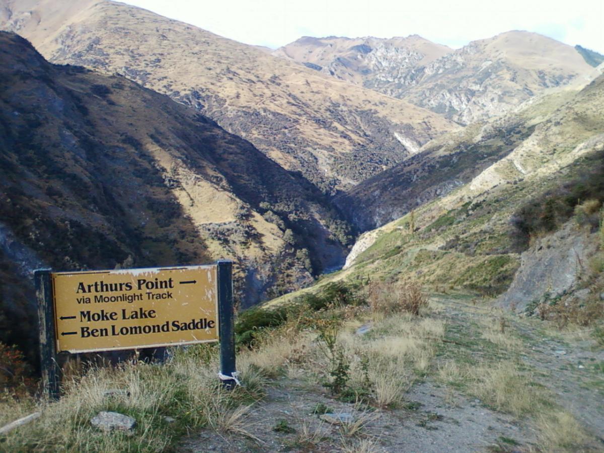 A scenic view of a mountainous landscape features a weathered wooden sign indicating directions to Arthurs Point via Moonlight Track, as well as Moke Lake and Ben Lomond Saddle. The surroundings include steep hills with patches of green and dry grass under a clear sky. Moonlight Circuit mountain bike trail.