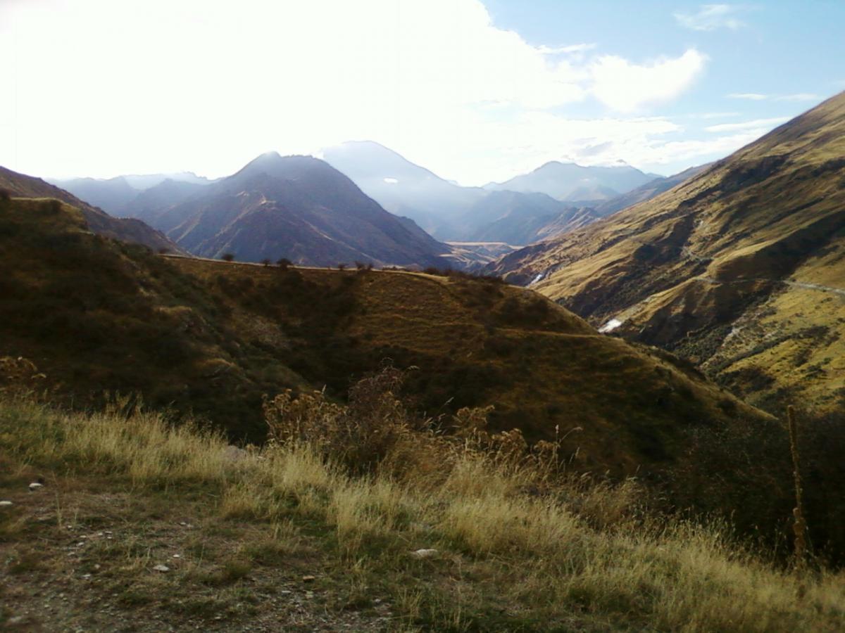 A scenic view of mountainous terrain featuring rolling hills, lush greenery, and a cloud-filled sky. The landscape is bathed in soft sunlight, highlighting the contours of the mountains in the distance. Moonlight Circuit mountain bike trail.