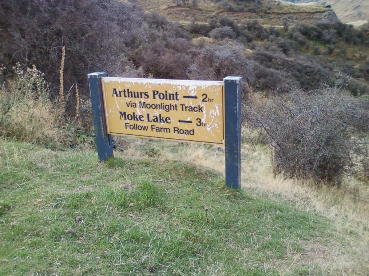 Signpost indicating directions and estimated travel times to Arthurs Point (2 hours via Moonlight Track) and Moke Lake (3 hours, follow Farm Road), set against a natural landscape with vegetation in the background. Moonlight Circuit mountain bike trail.