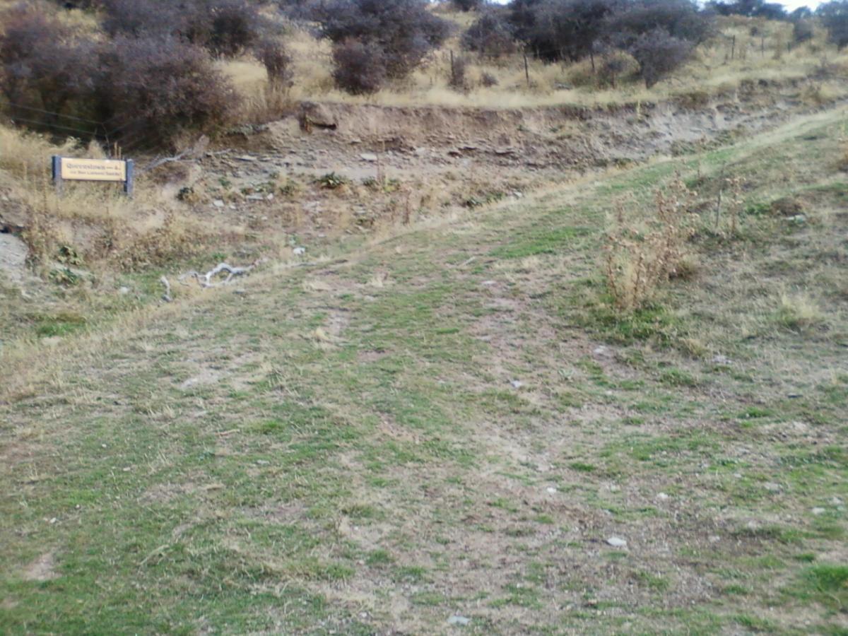A dirt path winding through a grassy area, with sparse vegetation and a sign on the left side indicating the name of a location or trail. The landscape is hilly and barren, with dry bushes in the background. Moonlight Circuit mountain bike trail.