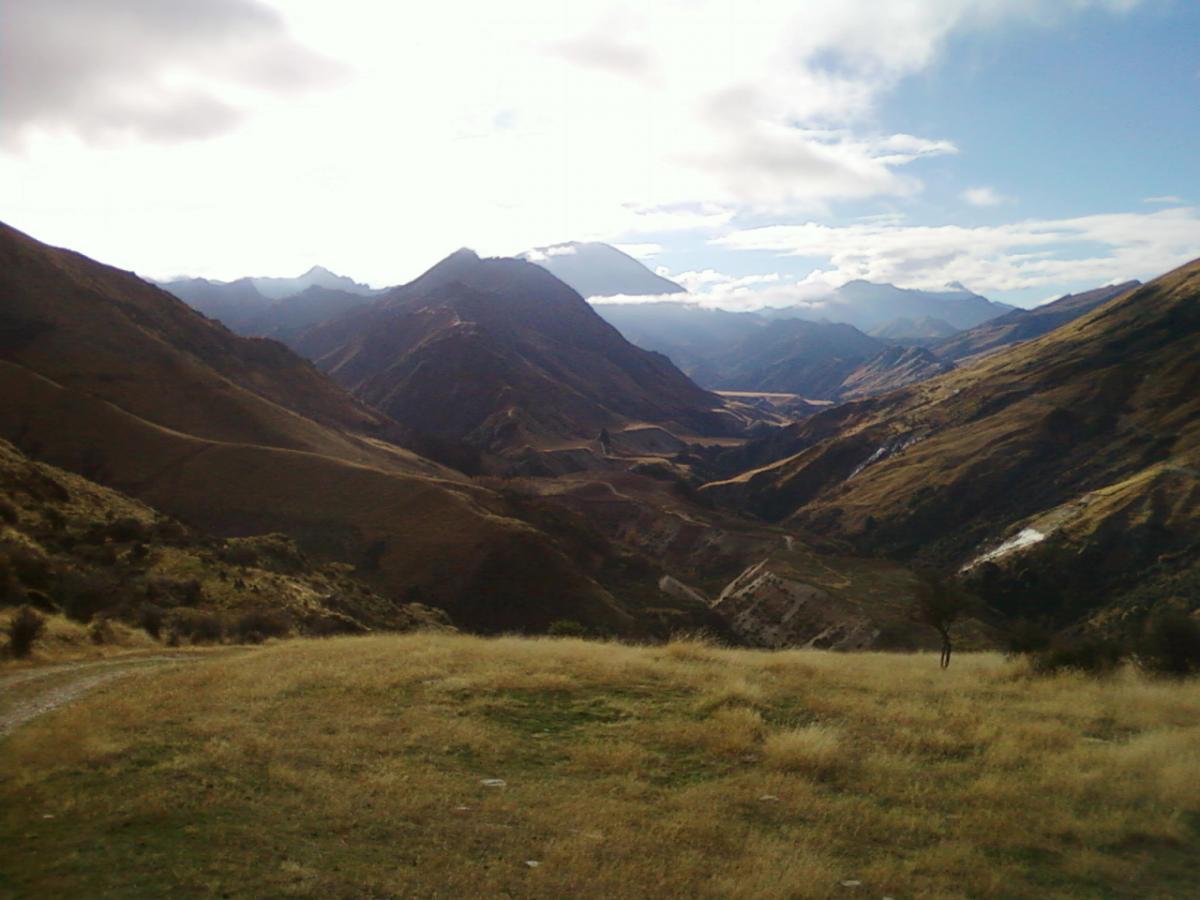 A panoramic view of a mountainous landscape featuring rolling hills and steep valleys under a partly cloudy sky. The foreground shows grassy terrain, while the background displays a series of mountains with peaks partially obscured by clouds. Moonlight Circuit mountain bike trail.