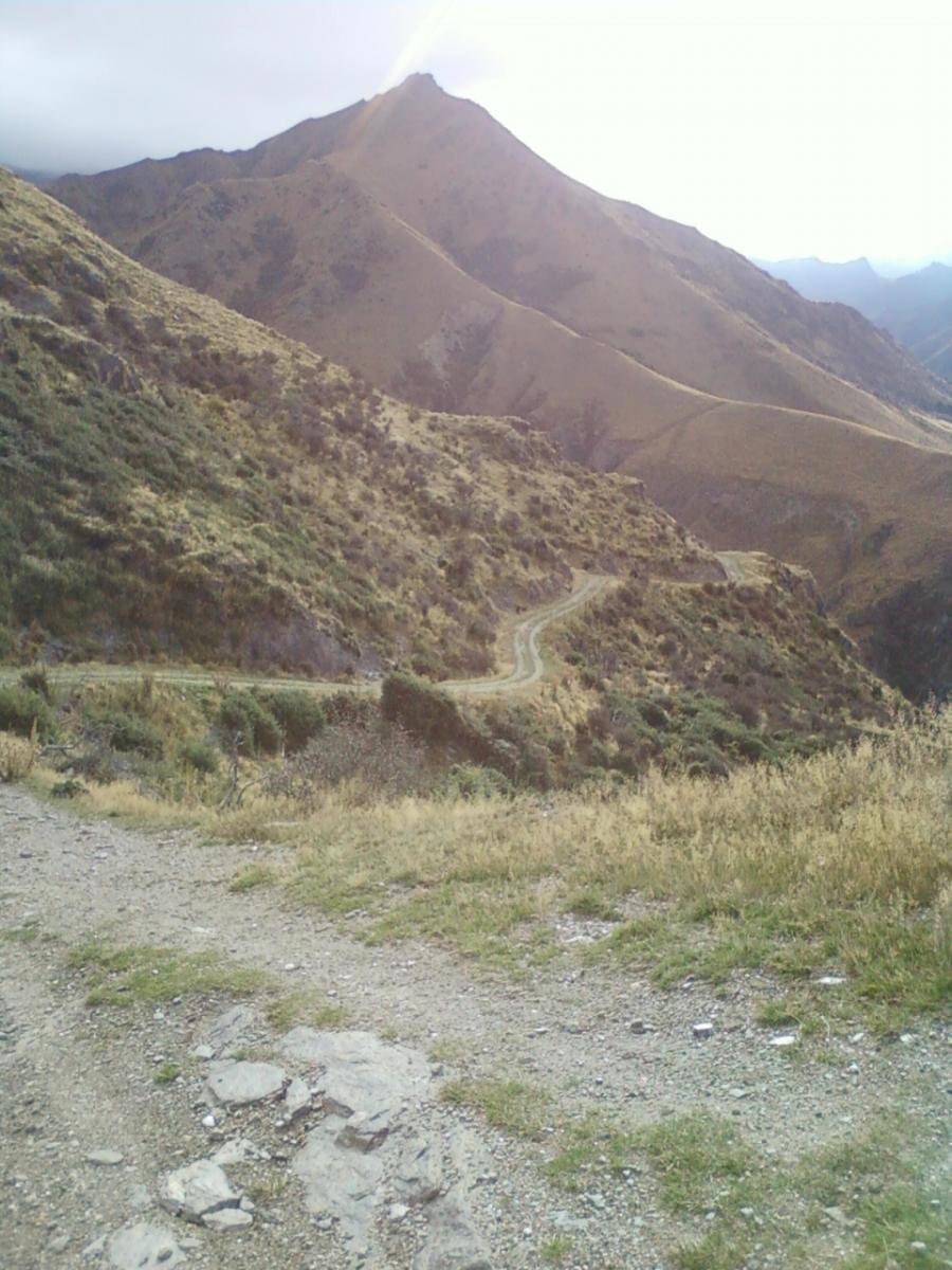 A scenic view of a mountainside with a winding dirt path leading through grassy terrain. The landscape features steep, dry hillsides and a cloudy sky, suggesting a cool, overcast day. The foreground includes rocky outcrops and sparse vegetation. Moonlight Circuit mountain bike trail.